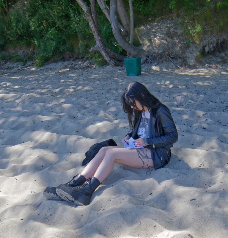 17 - Girl with tatoo sitting on a sandy beach, wearing a black leather jacket and black combat boots